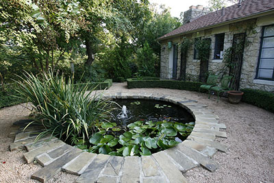 A small round pond full of green plants like lily pads and ornamental grasses plus a small bubbling fountain, in the center of a circular paved area in front of a shady home.
