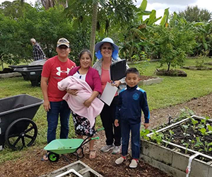 A woman standing and smiling in a garden alongside a family of a man, woman holding a baby wrapped ina pink blanket, and a young boy.