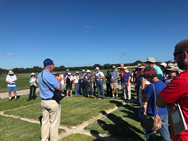 Man speaking to group out in a turfgrass field with squares of turf under foot and a bright blue sky overhead